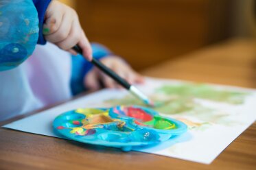 Vergrößerungsansichten für Bild: Kind mit Malschürze malt mit einem Pinsel, vor ihm ein Blattpapier und eine Farbmischpalette auf einem Tisch liegend 