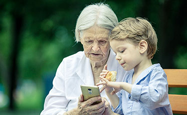 Vergrößerungsansichten für Bild: Nahaufnahme: Eine ältere Frau sitzt mit ihrem Enkel auf einer Bank und beide schauen sich etwas auf einem Smartphone an